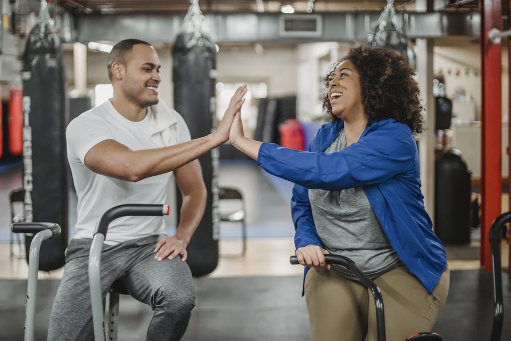 Happy plump African American female trainee and muscular smiling coach clapping each others hands while sitting on stationary bicycles in modern gym